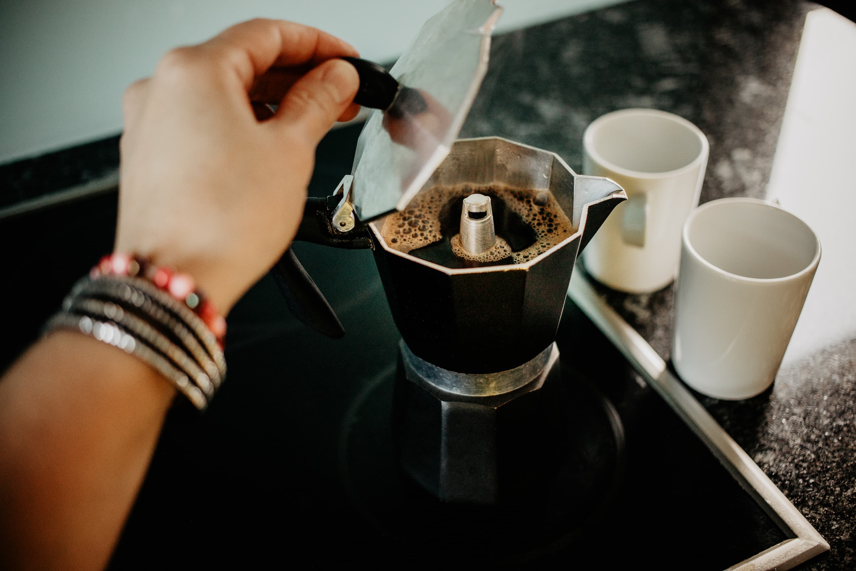 Hand öffnet Espressokocher auf Herd, frischer Kaffee steigt auf, zwei weiße Tassen daneben.