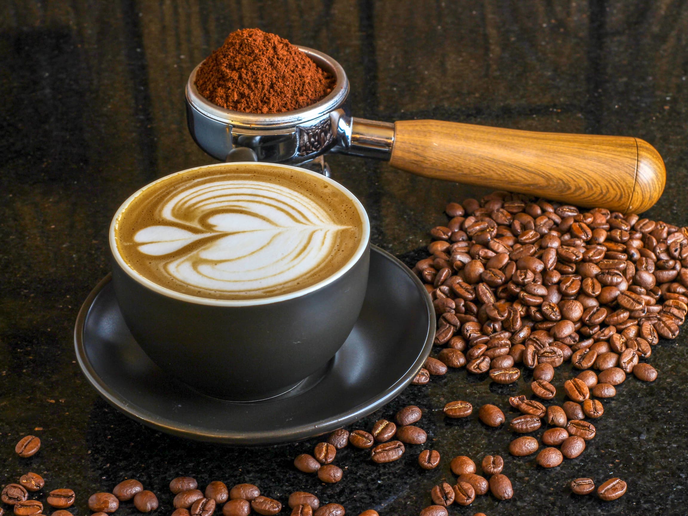 Schwarze Tasse mit Latte Art, daneben Kaffeebohnen und Siebträger mit Kaffeepulver.