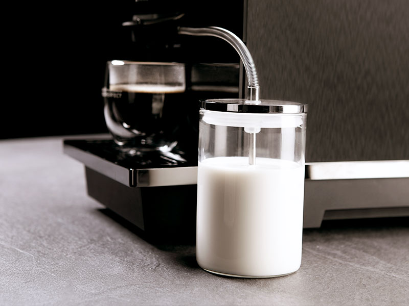 Transparenter Milchbehälter mit Schlauchanschluss an einem MP10 Kaffeevollautomaten auf grauer Arbeitsfläche.