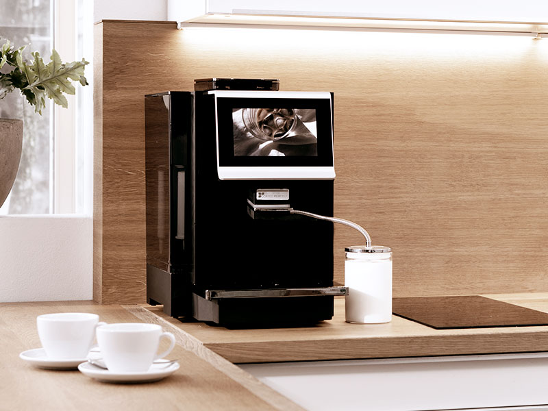 Schwarzer MP10 Kaffeevollautomat mit angeschlossenem Milchbehälter auf heller Küchenarbeitsfläche und zwei Tassen im Vordergrund.