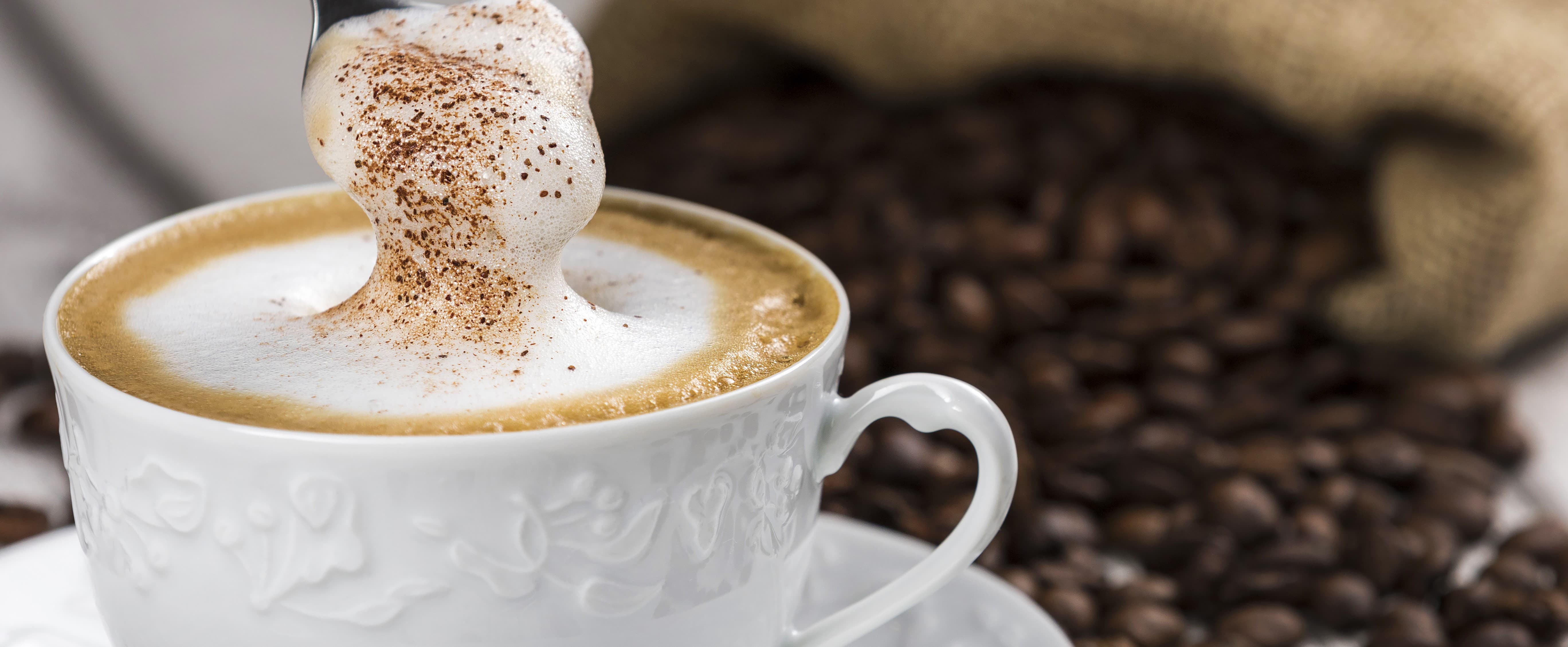 Weißes Porzellan mit Cappuccino, Milchschaum und Zimt, Kaffeebohnen im Hintergrund.