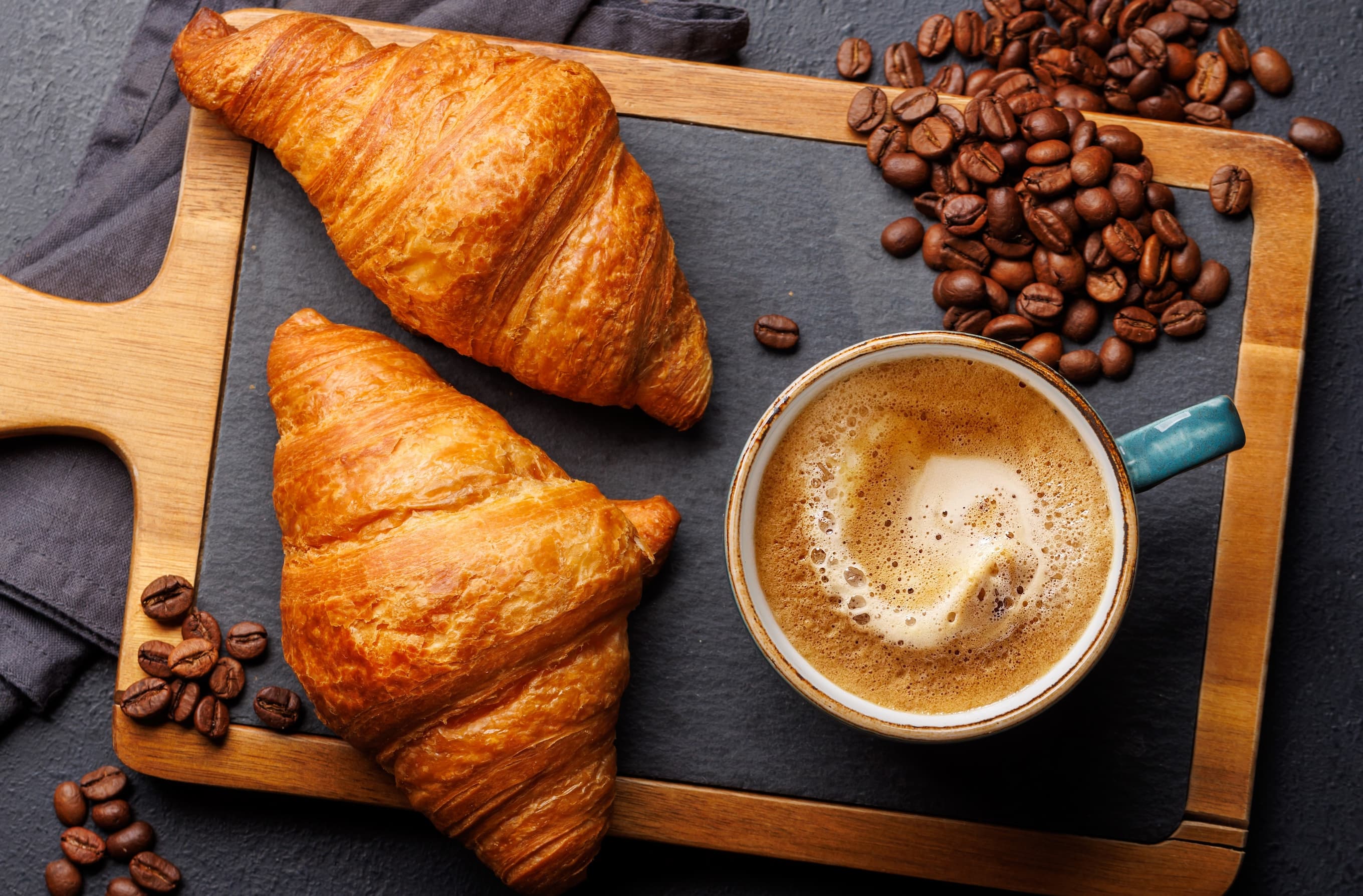 Zwei Croissants mit Tasse Kaffee und Kaffeebohnen auf Holzbrett – französisches Frühstück.