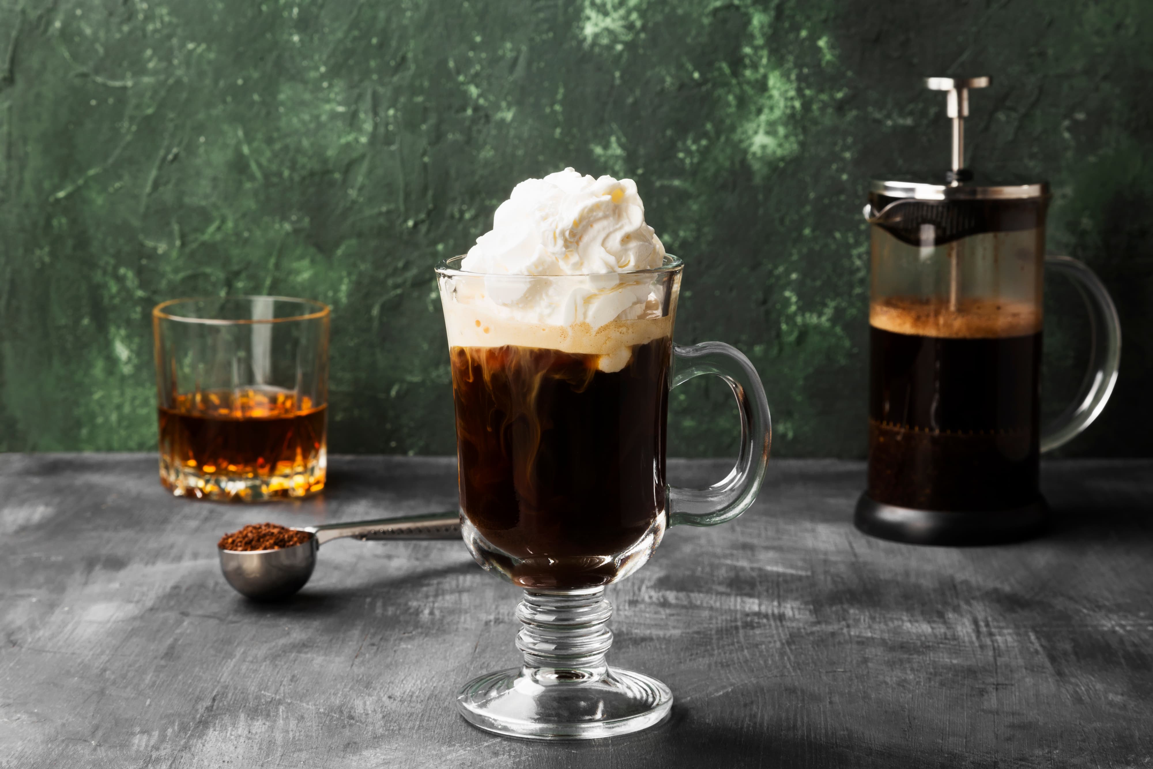 Glas Irish Coffee mit cremiger Schlagsahne auf grauer Tischoberflaeche, im Hintergrund Whiskeyglas und French Press.
