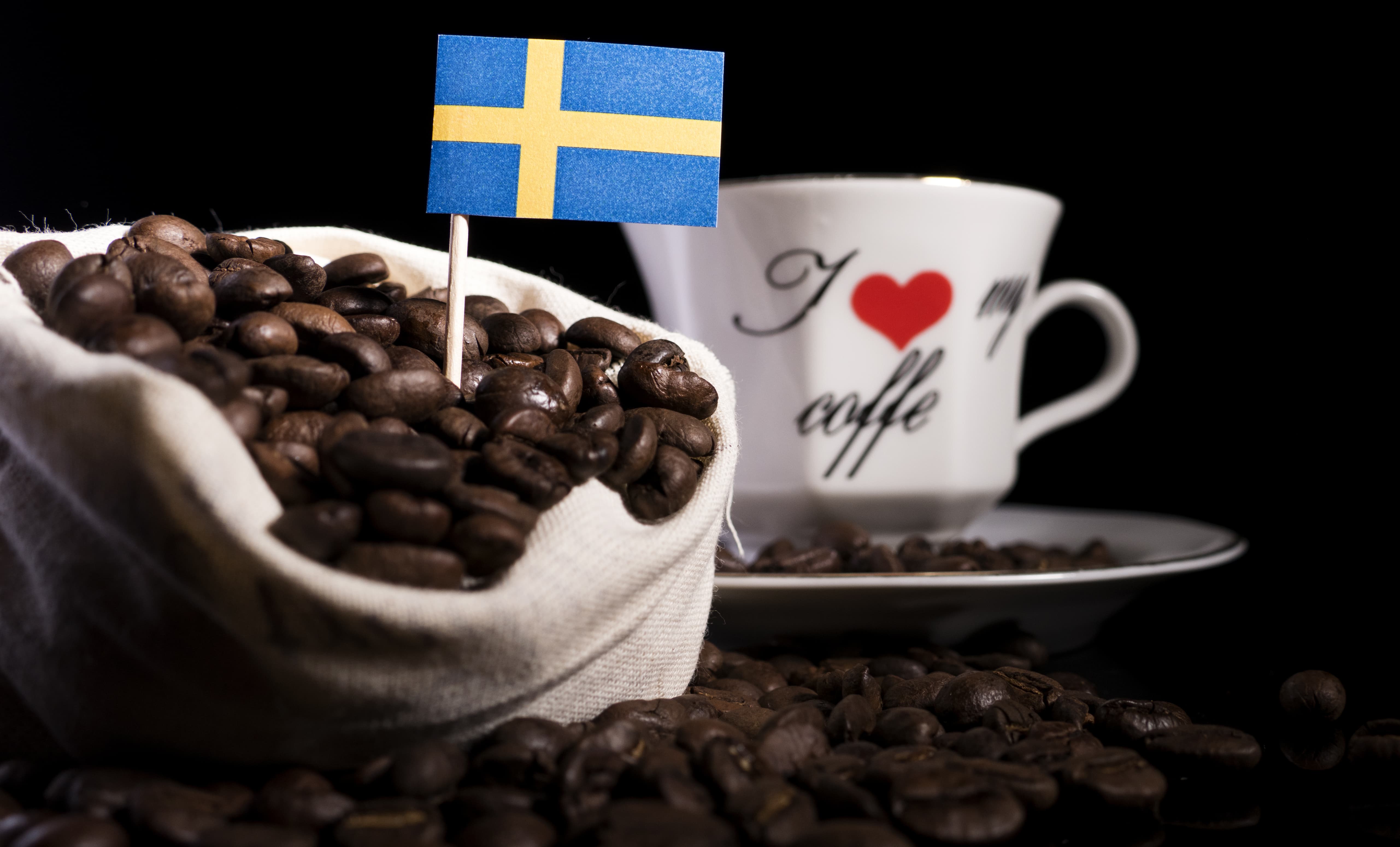 Sack voller Kaffeebohnen mit schwedischer Flagge, im Hintergrund eine weiße Tasse mit Aufschrift.