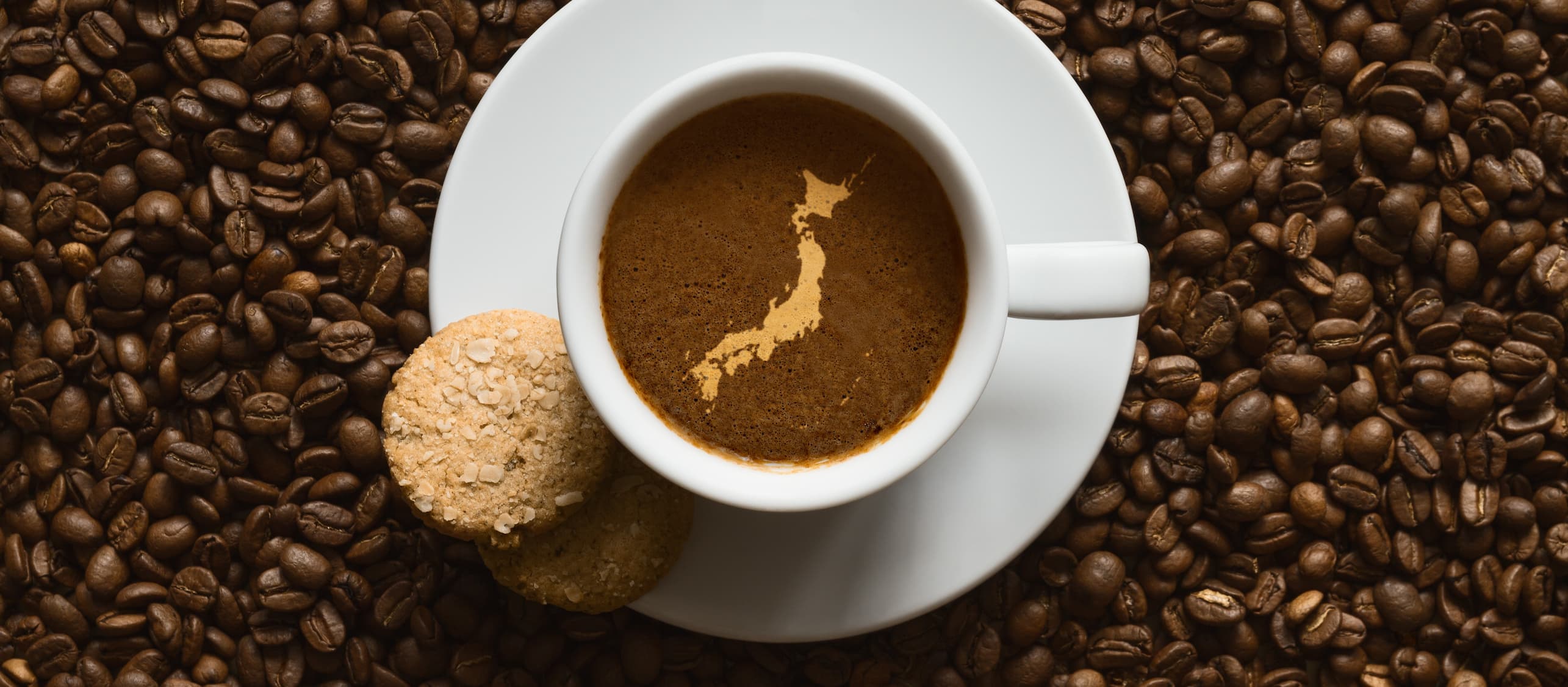 Tasse Kaffee mit Schaum in Form der Japan-Landkarte, umgeben von Kaffeebohnen und Keksen.