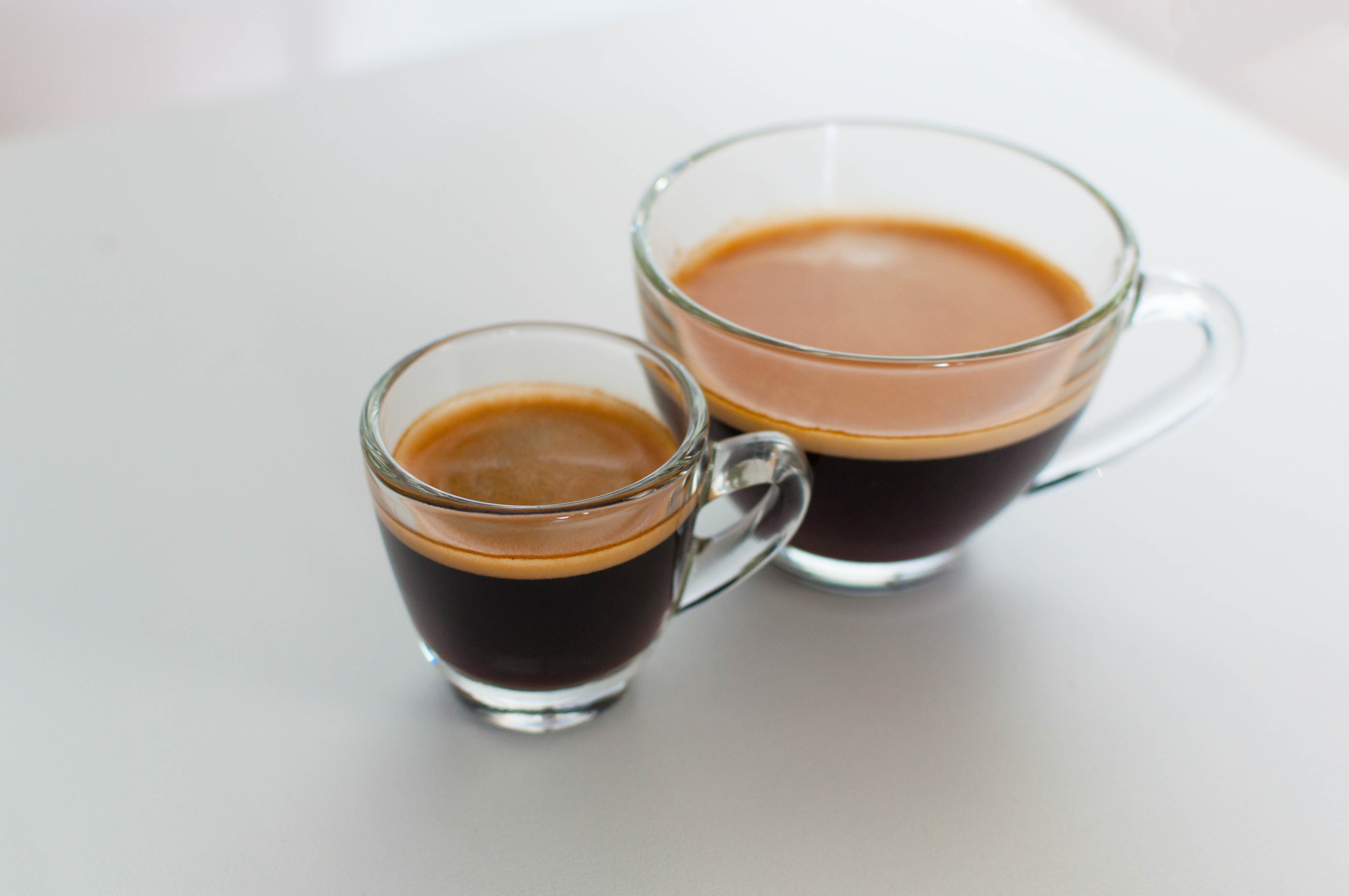 Zwei Glastassen mit Espresso und Americano auf hellem Untergrund, sichtbar unterschiedliche Fuellmengen und Crema.