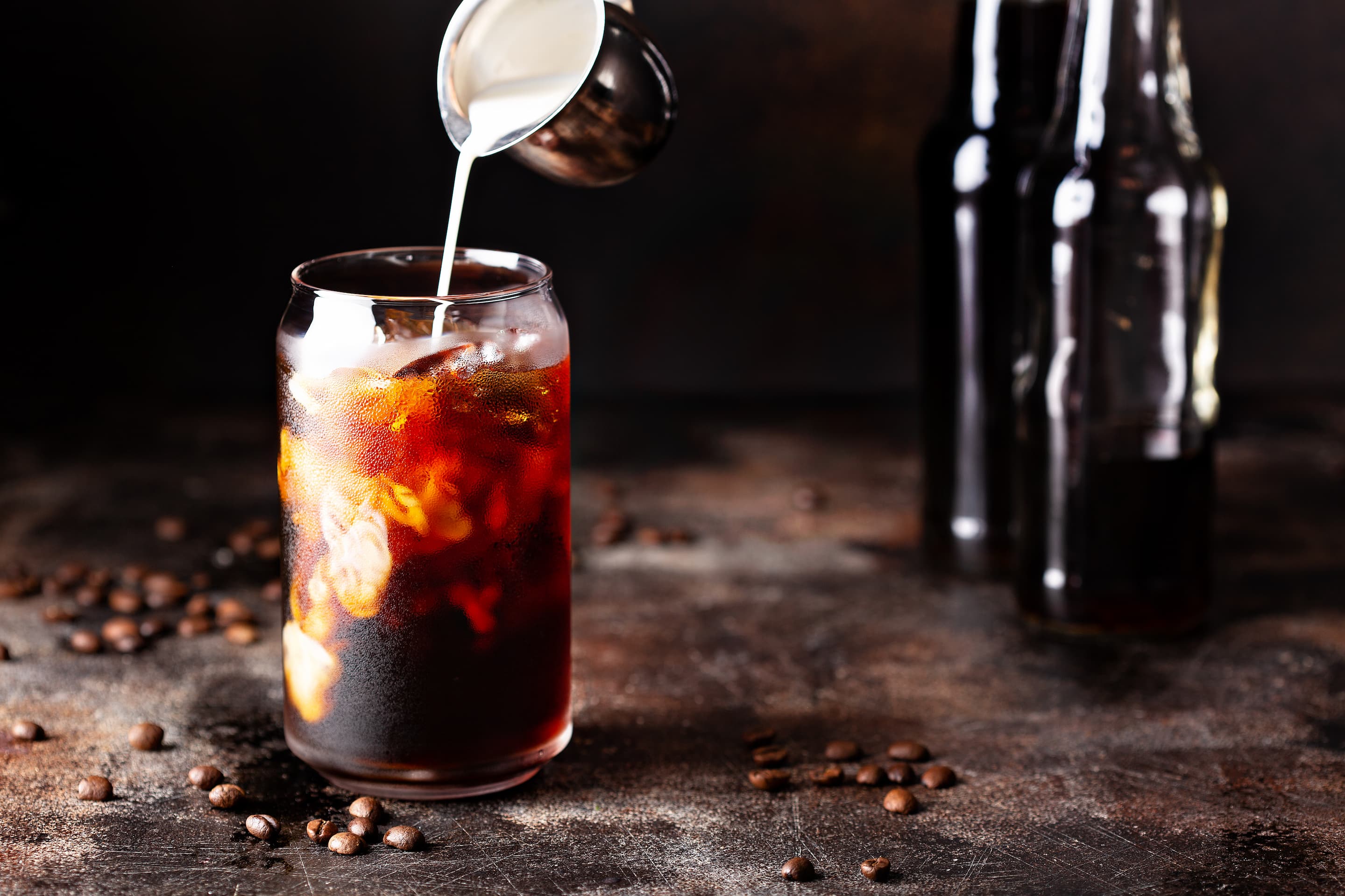 Glas Cold Brew Coffee mit Eiswuerfeln und Milch, daneben dunkle Flaschen im Hintergrund.
