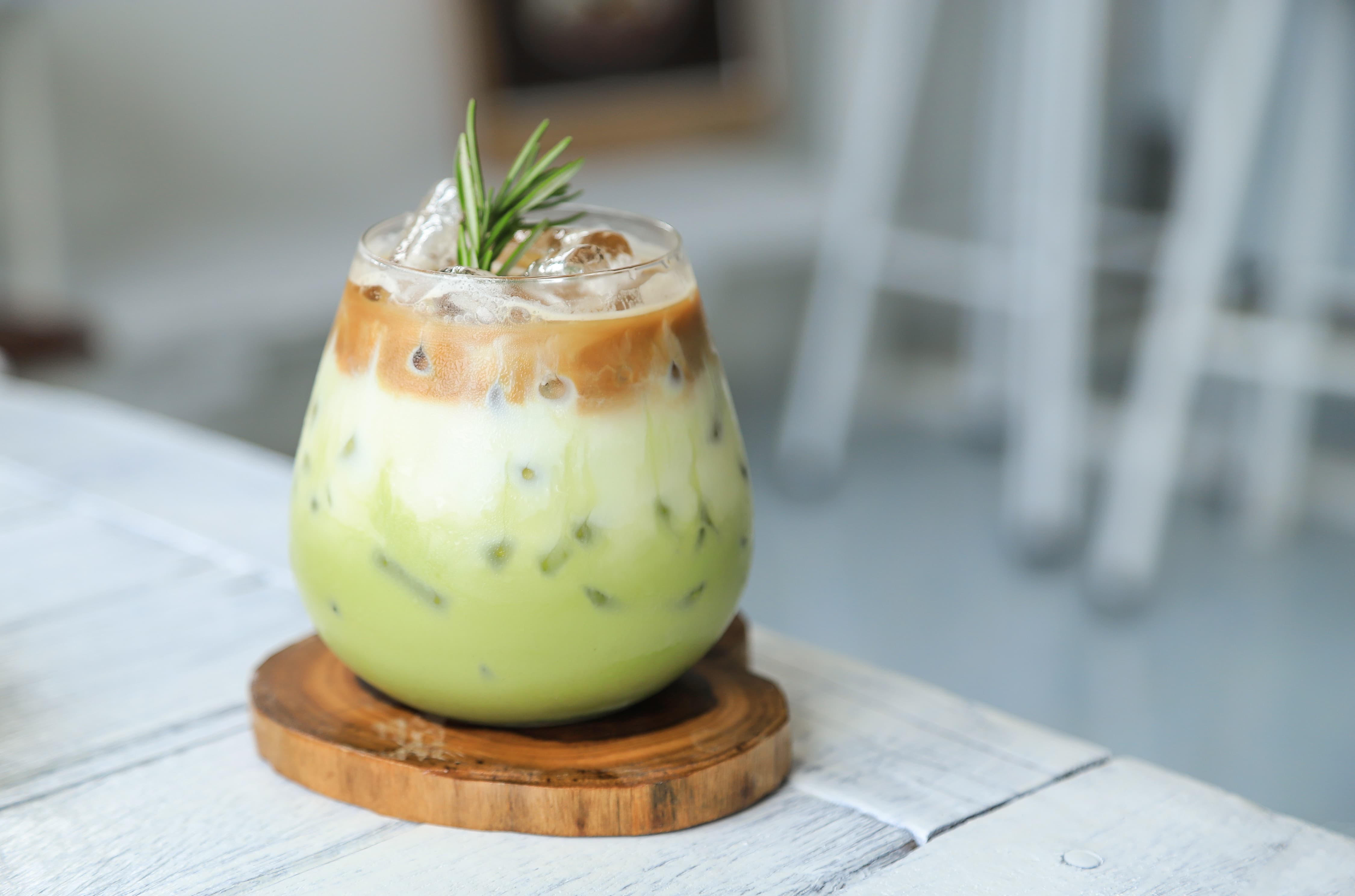 Glas mit Iced Matcha Latte, Schichten aus grünem Tee, Milch und Kaffee, dekoriert mit Rosmarin.
