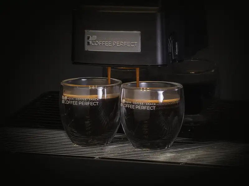Zwei Tassen Kaffee unter einer laufenden Kaffeemaschine Zwei Tassen Kaffee unter einer laufenden Kaffeemaschine